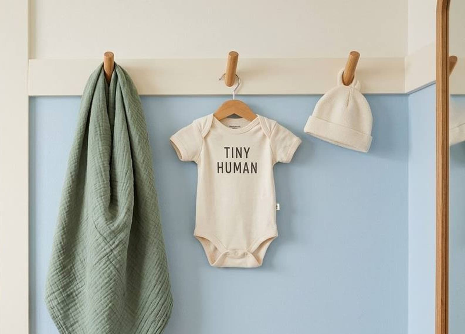 The sage Burp Bandana hanging from a wooden peg rail in a nursery, beside a TINY HUMAN onesie and a knotted baby hat