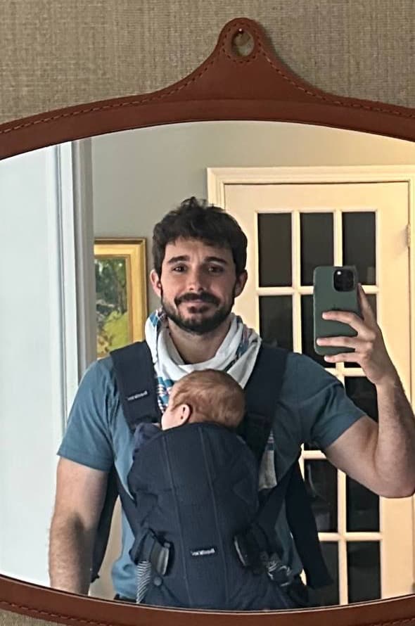 Mirror selfie of Zack with baby Asher asleep in a front carrier, a hospital blanket knotted around his neck as a makeshift bandana