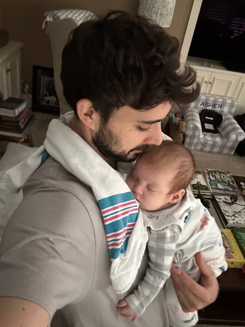 Zack holding his newborn son Asher against his chest and kissing the top of his head, a striped hospital blanket draped around his neck and down over his shoulder