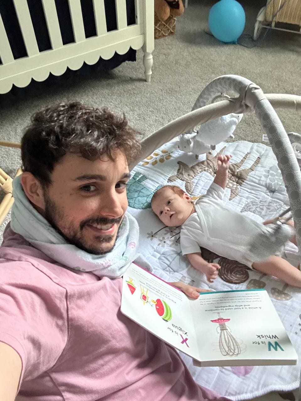 Zack lying on the nursery floor reading an alphabet book to baby Asher on a play mat, grinning, still wearing the hospital blanket around his neck